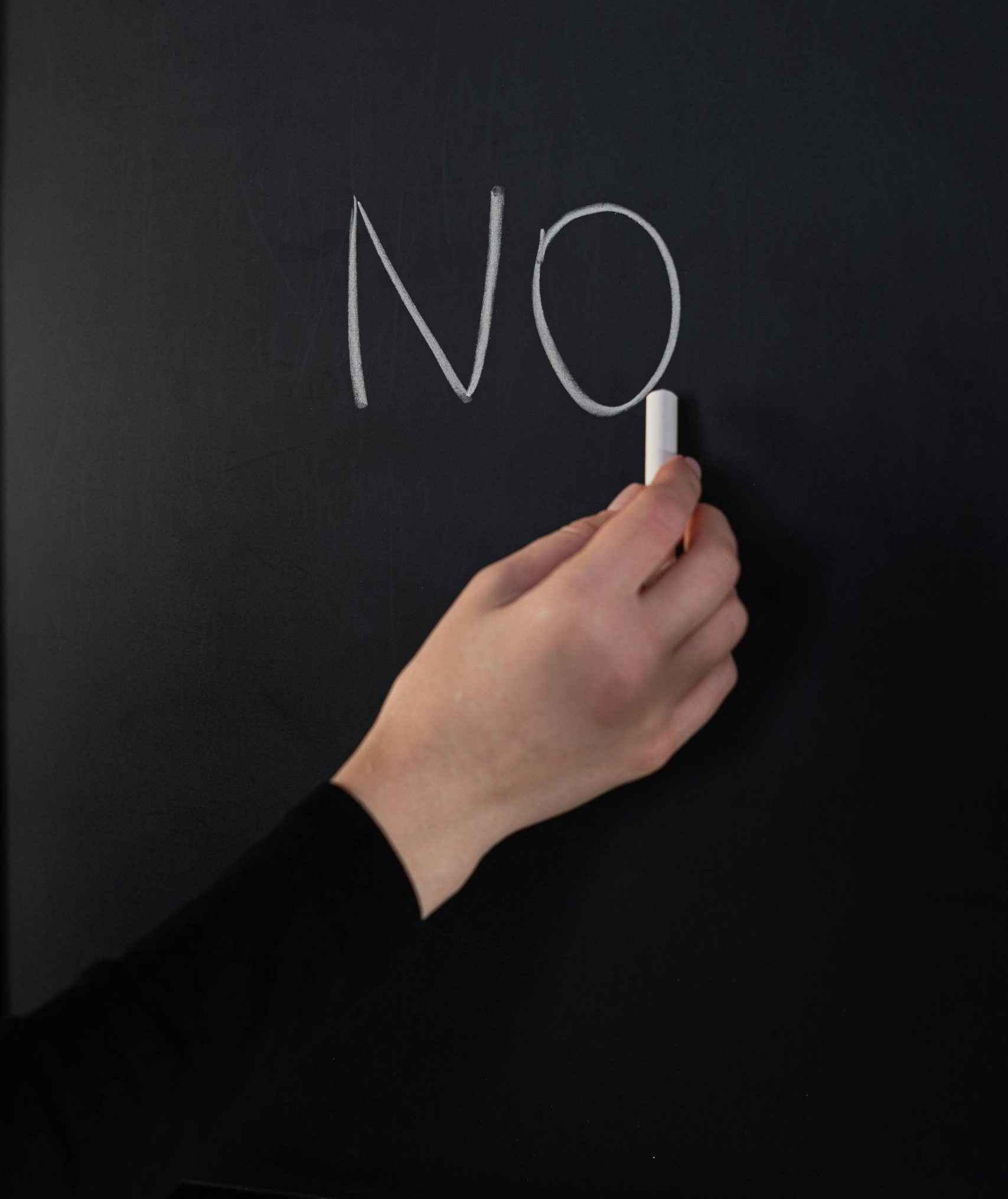 Close-up of a hand writing 'NO' on a chalkboard using white chalk.